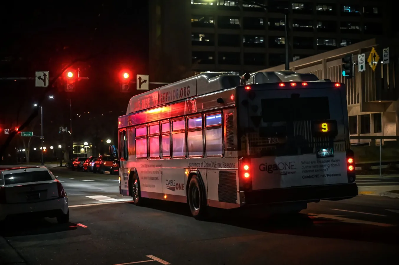 A city bus stopped at a traffic light during the night in an urban downtown setting.