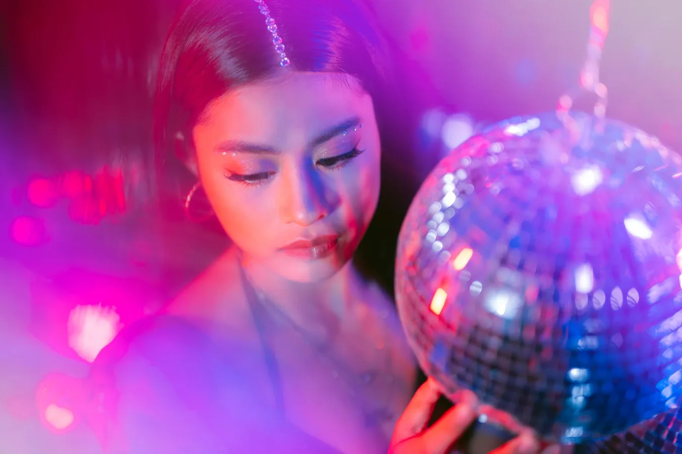 Close-up of a woman with glitter makeup holding a disco ball under vibrant neon lights.