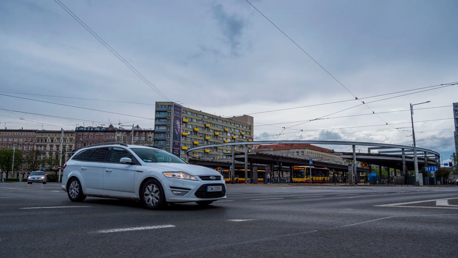 City street with a white car, buses, and modern buildings under a cloudy sky.