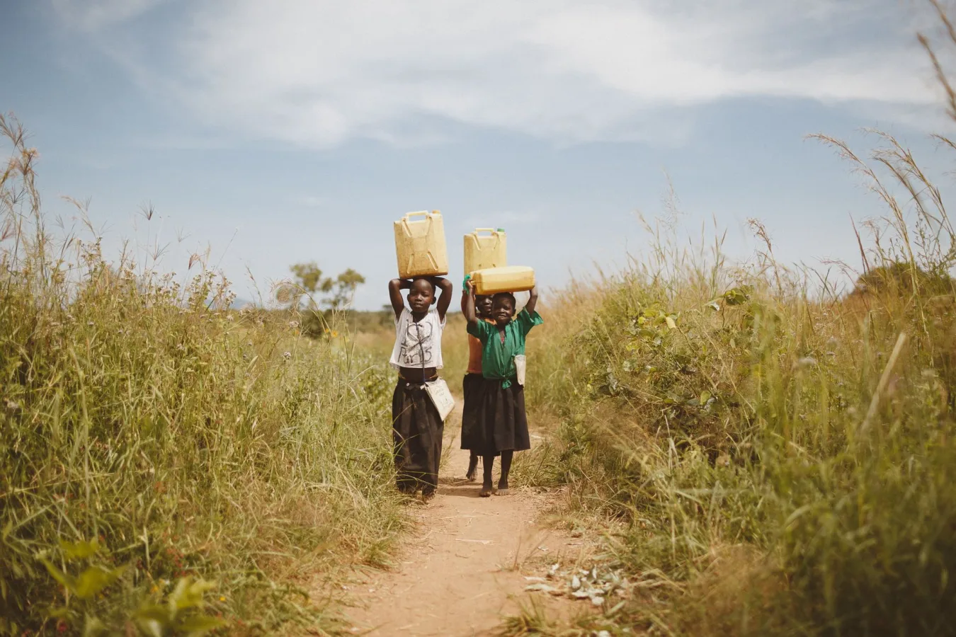 Two children walking through a rural pathway carrying water containers on their heads in K